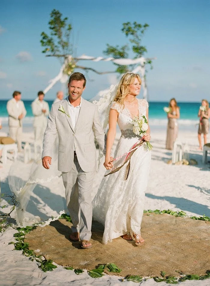 relaxed boho wedding at the beach, happy bride and groom, dressed in a white retro lace gown, and pale gray suit, walking while holding hands
