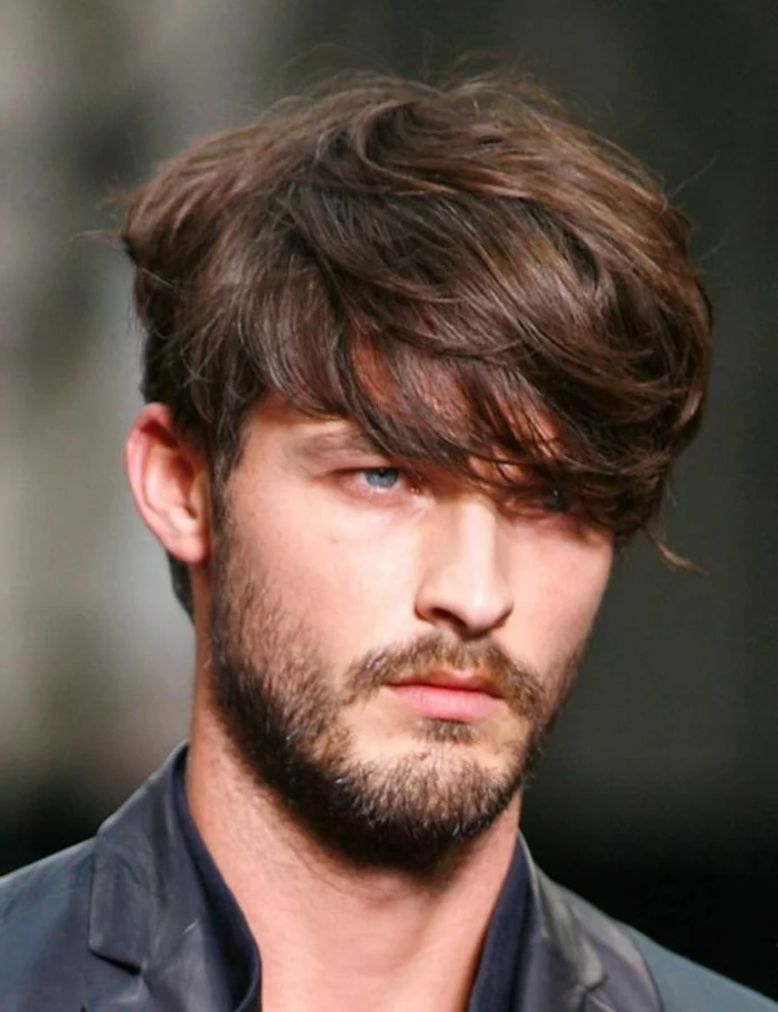 bangs falling over one eye, brunette textured and layered haircut, worn by blue-eyed man, with short beard and mustache