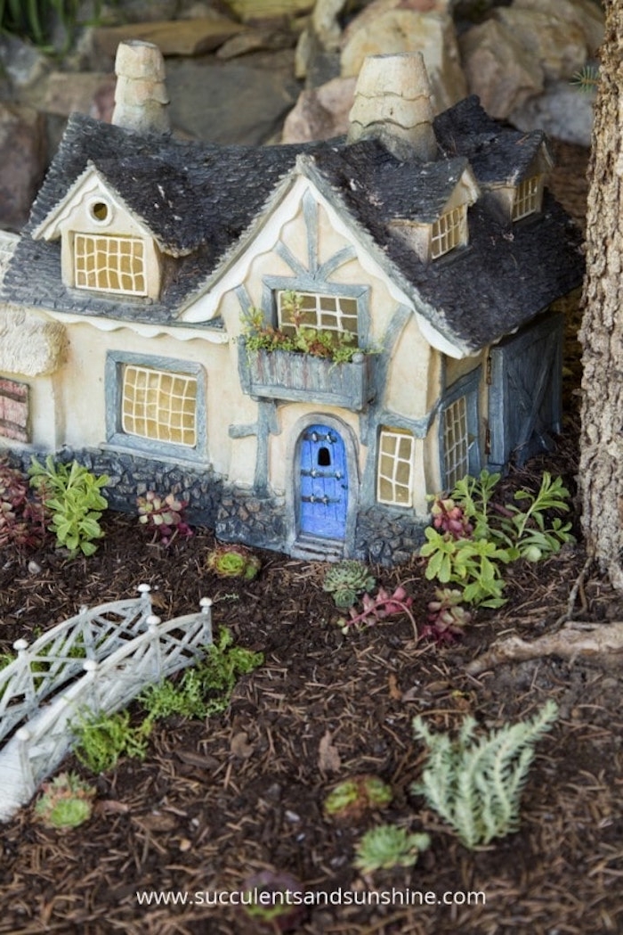 dark blue roof, light blue beams and window frames, and a bright blue door, on a miniature fairy house ornament, placed on a forest floor, near white toy bridge