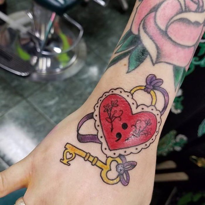 several tattoos on a slim arm, large pink rose with green leaves, heart-shaped lock in red and white, with a black semicolon keyhole, semicolon meaning, yellow key and purple ribbon