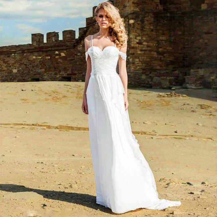 antique castle wall, on a sandy seashore, slim blonde woman with curly hair, wearing white maxi dress, with straps and slouched sleeve details