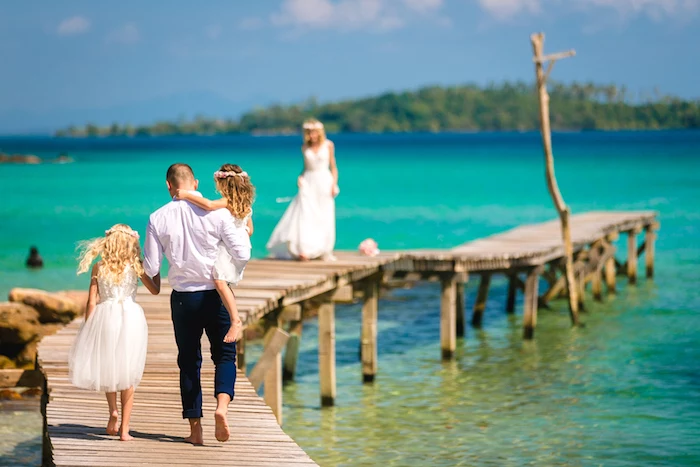 beach wedding venues, tropical sea in azure blue, man and two young girls, walking on a boardwalk, towards a blonde woman, in a long white gown