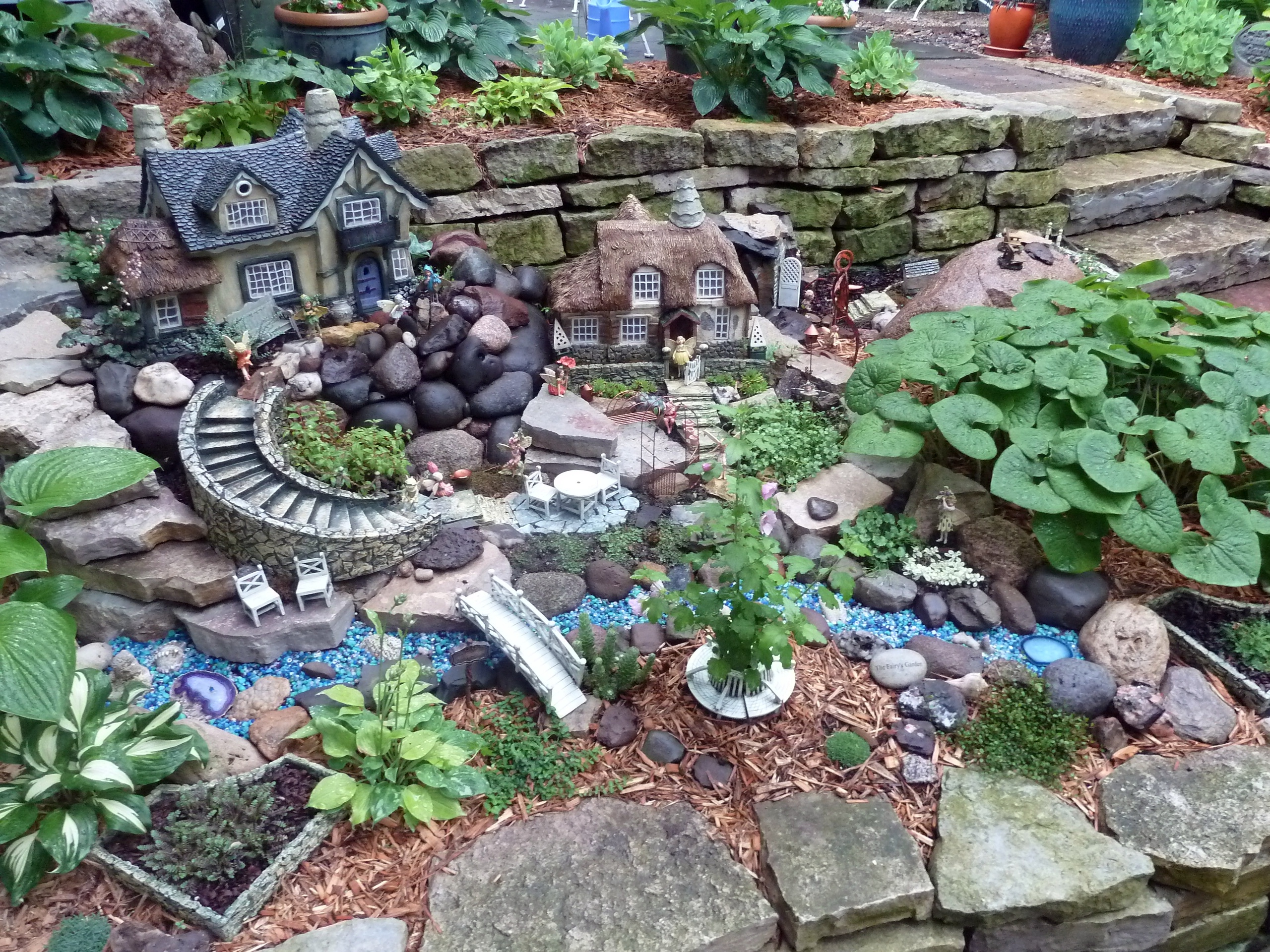 elaborate garden set up, including two fairy houses, a tiny winding staircase, several figurines and accessories, how to make a fairy garden, different kinds of plants 