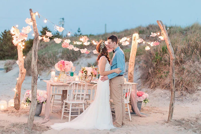 pink flowers in different tones, in bouquets and garlands, on and near a set white rustic table, with matching chairs, florida destination weddings, young couple kissing