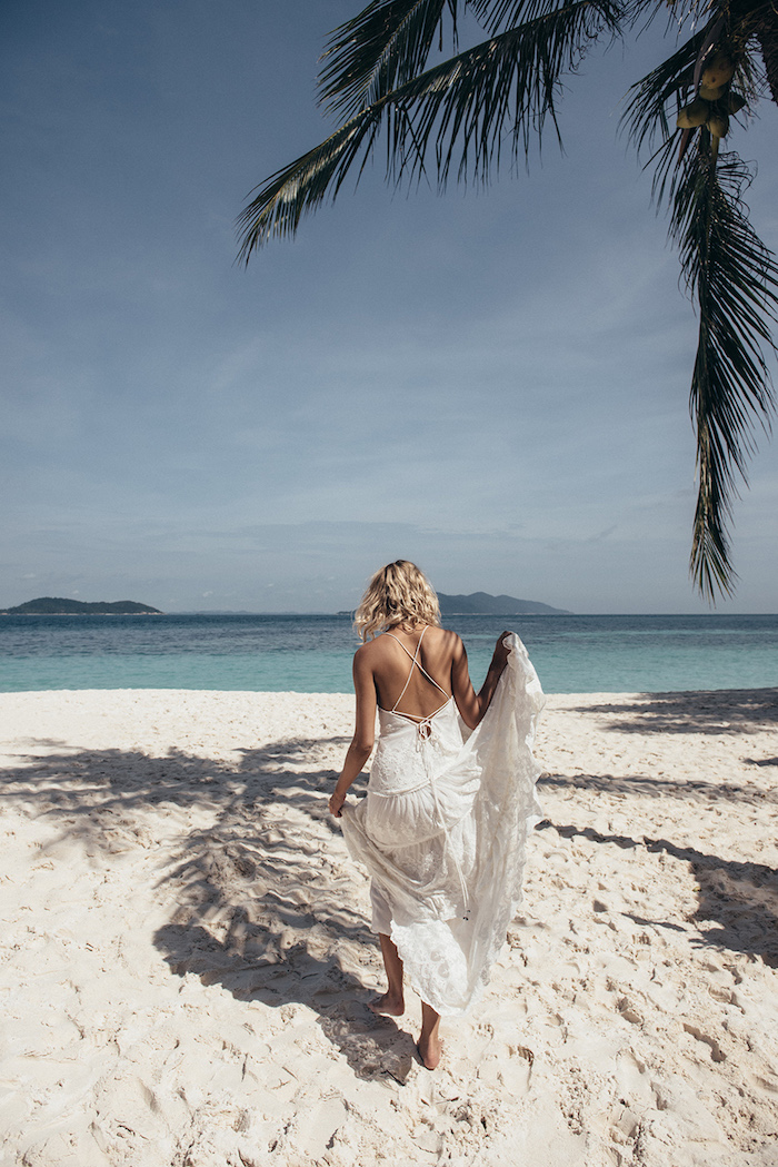palm tree on a sandy beach, near an azure blue sea, curly blonde woman, walking towards the water, holding the skirt of her white, embroidered boho gown, beach wedding dresses