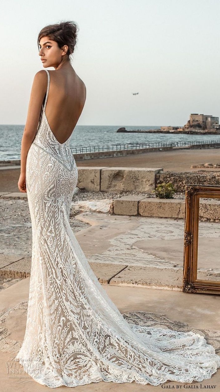 backless white and nude mermaid-style, lace beach wedding dress, worn by tanned young woman, with brunette hair in a messy bun, standing near the sea