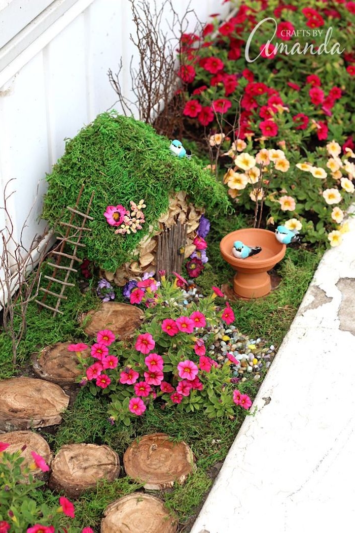 birdbath of a miniature size, placed near a diy fairy house, with moss-covered roof, and tiny ladder, in a garden with pink, red and yellow flowers