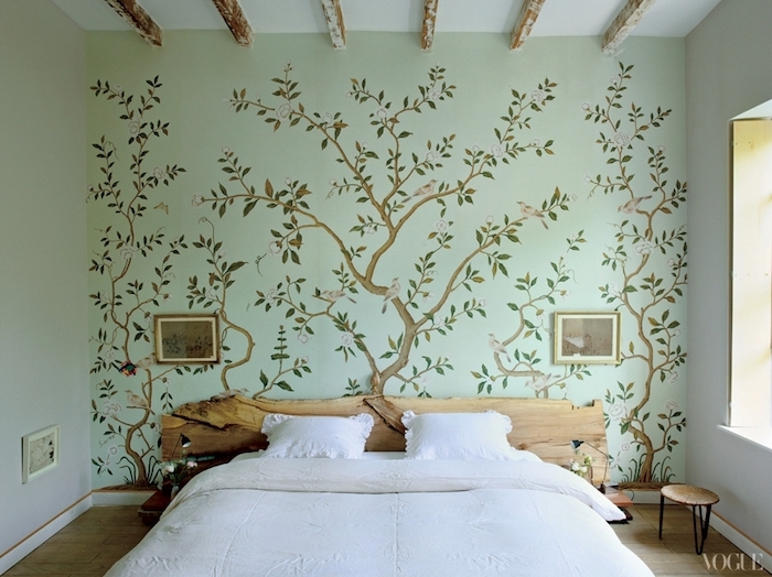antique-style mural, with thin blossoming trees, with green leaves, and small gray birds, in bedroom with wooden ceiling beams, double bed with headboard, made from a piece of natural wood