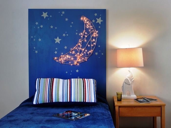 string lights attached to a blue board, forming a glowing moon shape, pale yellow star shapes, bedroom wall decor, single bed with blue velvety bedding, and a striped pillow