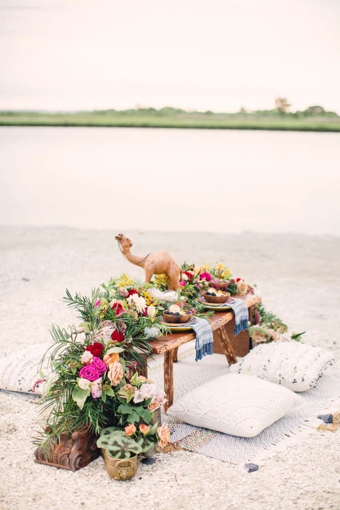 wooden table covered in many flowers, placed on a pale rug near several cushions, with a camel decoration, and plates containing fruit