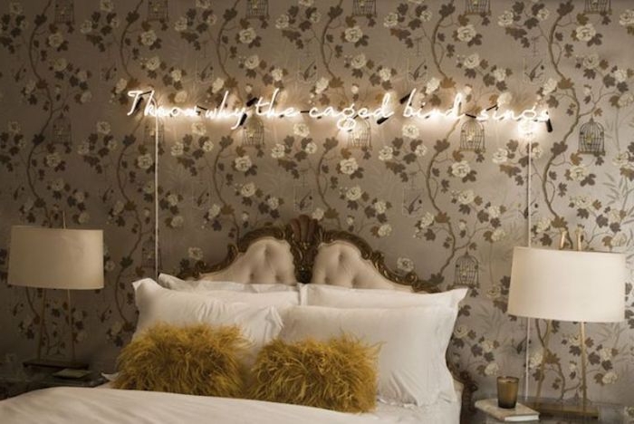 baroque style headboard, in white and gray, on plain white bed, with two fluffy yellow cushions, bedroom decorating ideas, floral wallpaper with neon writing