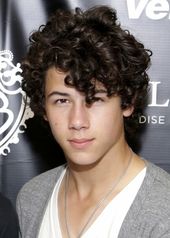 one of the jonas brothers, nick jonas with dark hair, many small curls, some falling over one of his eyes, curly hairstyles, grey cardigan and white top