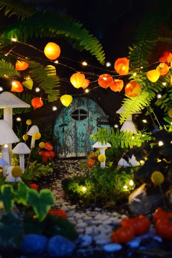 lanterns and tiny string lights, inside a night-time fairy garden, white mushroom figurines, pebbled path and various green plants, miniature teal-colored shabby door