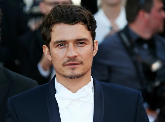 short curly hair, worn by Orlando Bloom, in white formal shirt, matching bowtie and black tuxedo, short beard and mustache