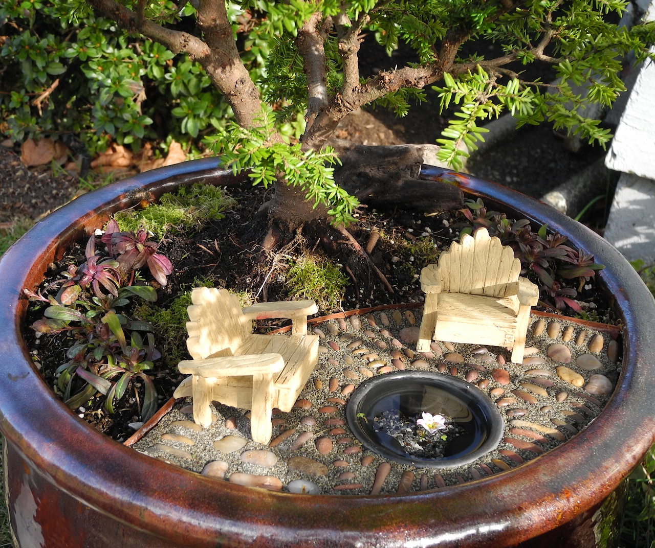 lounging chairs made of light wooden material, tiny figurines placed on pebbled surface, near miniature artificial pond, succulents and a bonsai tree