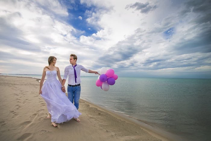 pale purple sleeveless wedding gown, worn by young smiling bride, holding hands with a groom in a matching shirt, and a dark purple tie, combined with jeans, beach weddings in florida, sandy shore and sea