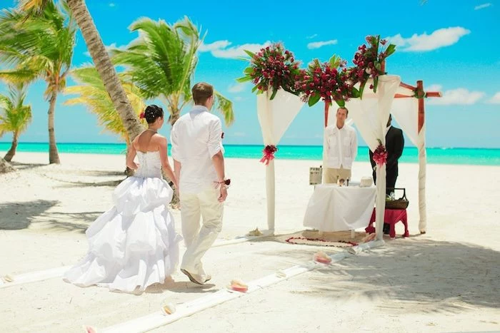 bali nuptials ceremony, white sandy beach, azure sea and blue sky, couple walking towards an altar, decorated with white fabric and flowers, beach wedding venues, palm trees swaying in the wind