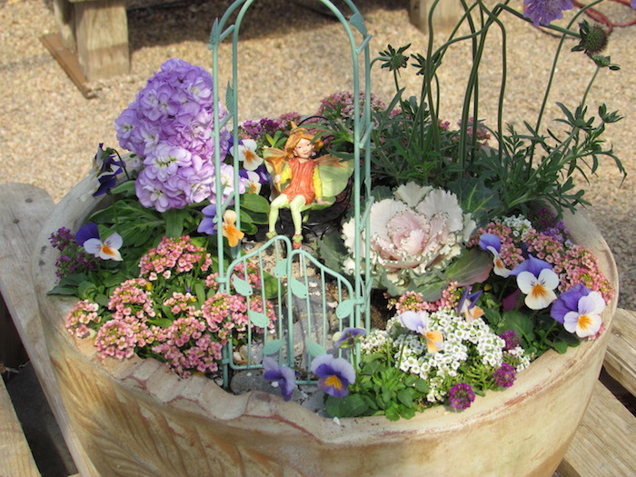 pale pink and purple flowers, with dashes of yellow, planted inside a large beige pot, decorated with a miniature blue wrought metal arch, with gates and a fairy figurine