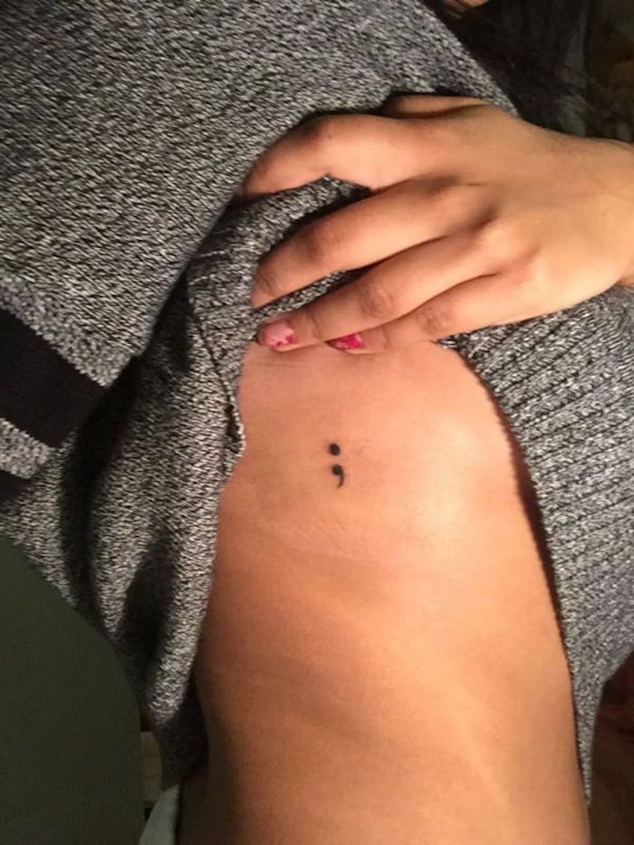 young woman holding up her salt and pepper gray jumper, to reveal a minimalistic black tattoo, of a semicolon, on her side, purpose of semicolon, chipped pink nail polish