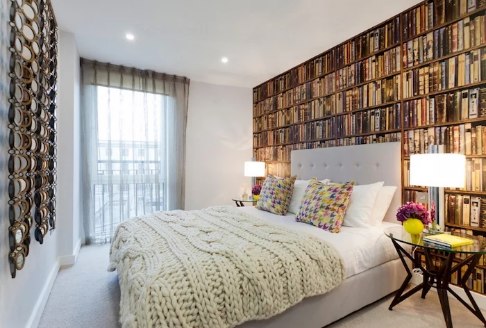 library photo wallpaper, with many different books, on a wall behind a pale gray double bed, master bedroom ideas, multi colored cushions, cream chunky knit throw
