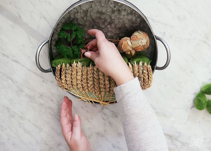 miniature deer figurine, placed near a tiny tree, small wooden fence, and 2 dried mushrooms, by two female hands, fairy garden with wheat stalks, inside metal cooking pot