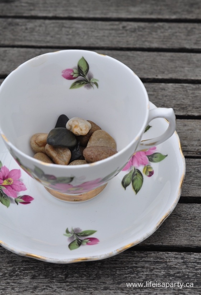 porcelain teacup in white, with a pink and green floral motive, and s matching saucer, half filled with pebbles in different colors, how to make a fairy garden