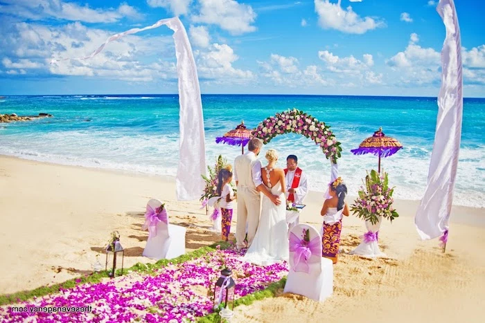 colorful wedding ceremony, on a beach with fine, pale yellow sand, and azure blue sea, purple and white flowers, decorating the aisle and the wedding arch, couple standing in front of a minister