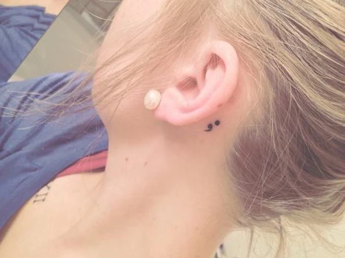 tiny black semicolon tattoo, behind a woman's ear, large pearl stud earring, blonde hair tied back, semicolon meaning and symbolism