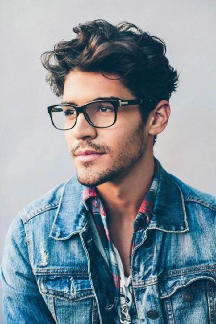 plaid shirt worn over a t-shirt, with a retro-style, ripped denim jacket, on man with brunette hair, styled in messy curls, hairstyles for short curly hair, stubble and glasses
