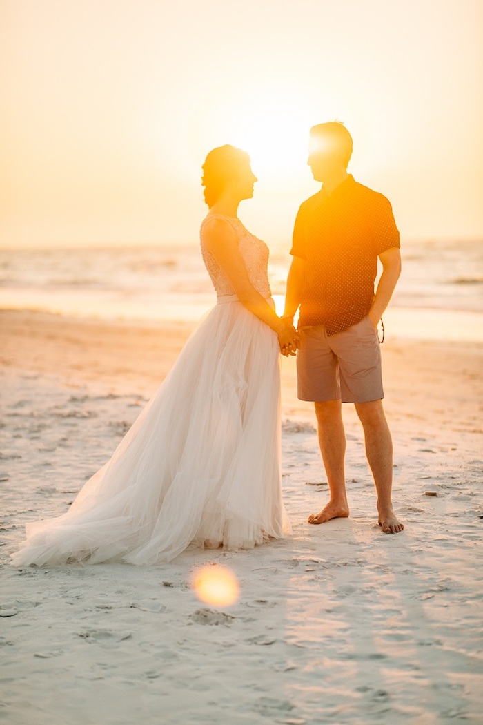 fine white sand, sea and a sunset, illuminating a couple holding hands, bride in long floaty tulle gown, groom in beige shorts, and a short-sleeved shirt, florida destination weddings