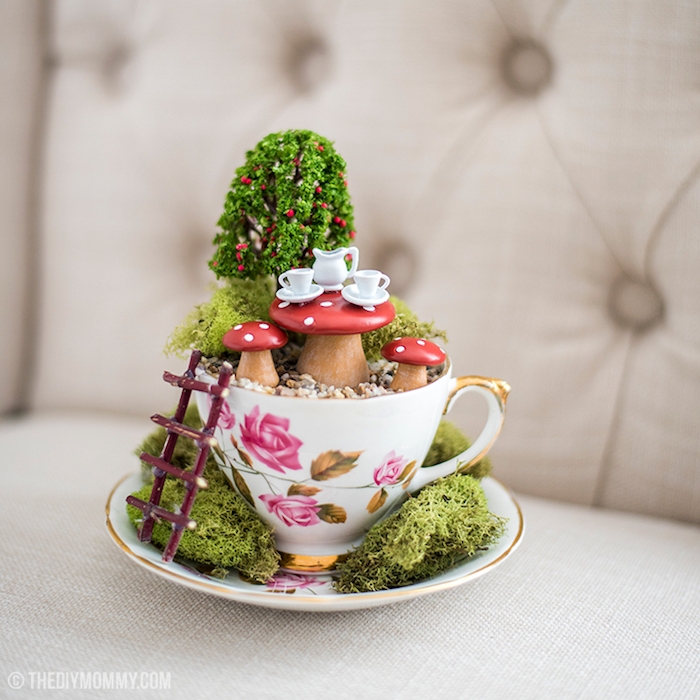 tiny white tea set, on a small mushroom table, with two matching chairs, placed in a small garden, how to make a fairy garden, with moss and a faux tree, inside a porcelain teacup