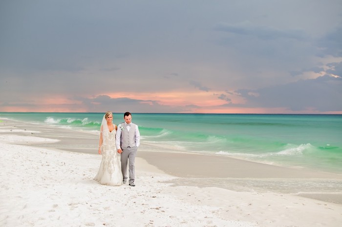 couple walking hand in hand, bride with embroidered off-white dress, and groom in grey two piece suit, sunset and white sand, beach wedding 