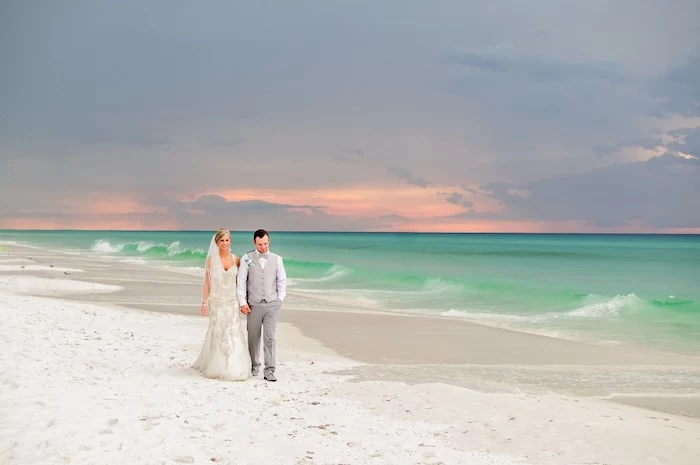 couple walking hand in hand, bride with embroidered off-white dress, and groom in grey two piece suit, sunset and white sand, beach wedding 