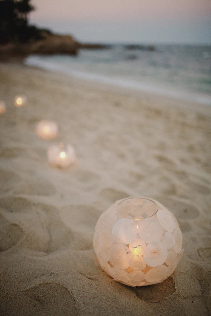several round glass lanterns, decorated with circles of cream-colored rice paper, containing lit candles, placed on fine beige sand, near the sea
