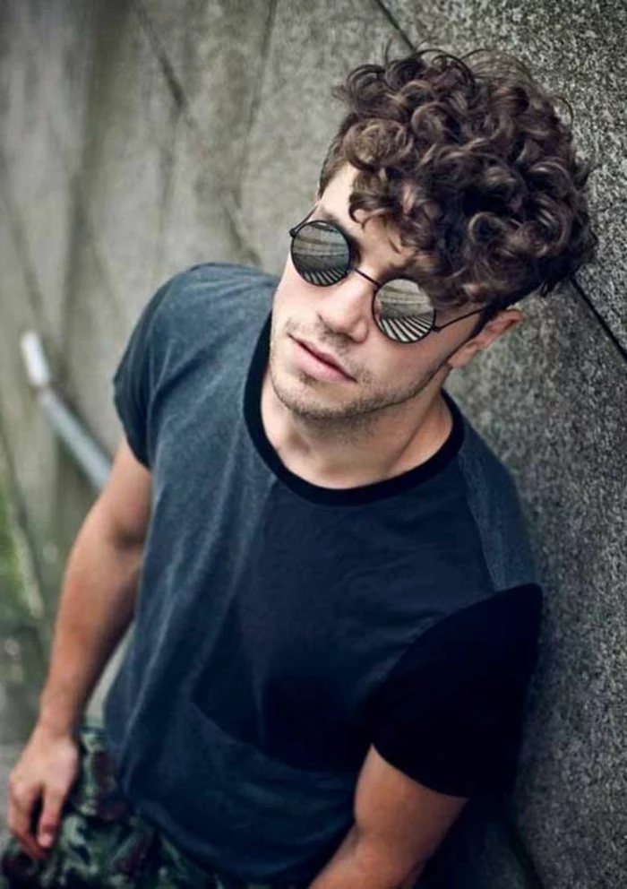 young man with curly brunette hair, wearing round reflective sunglasses, and a grey t-shirt, curly hairstyles, he is leaning on a gray wall