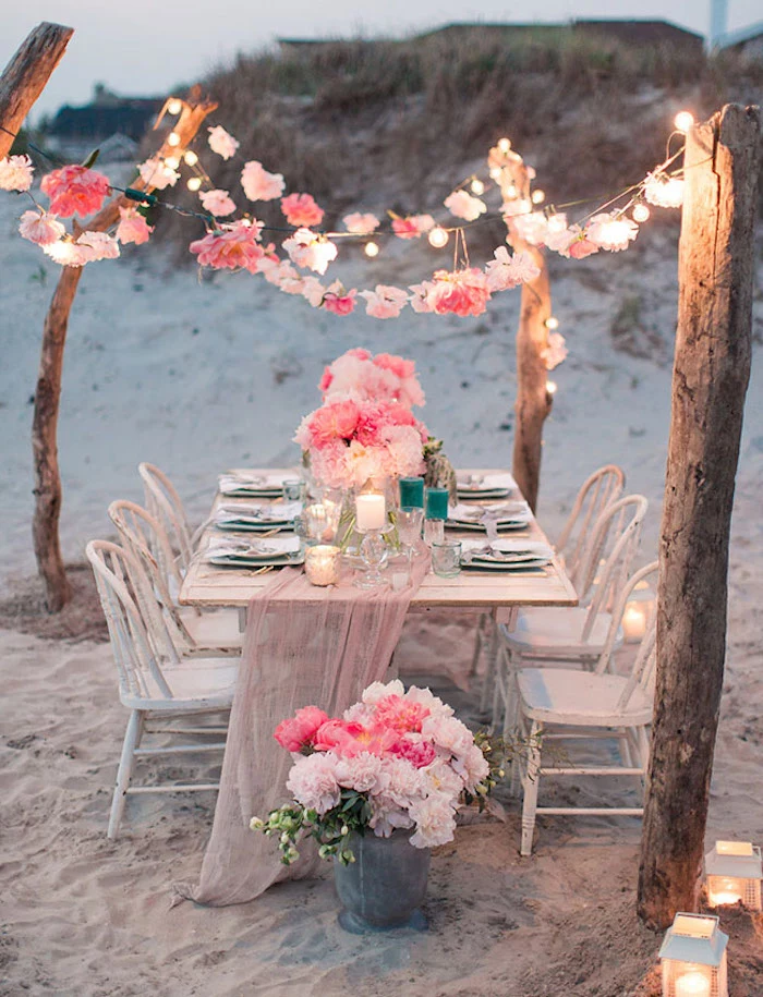 shabby chic white table, set up with plates, candles and pink flowers, six matching vintage chairs, on a white sandy beach, florida destination weddings, floral garlands and string lights