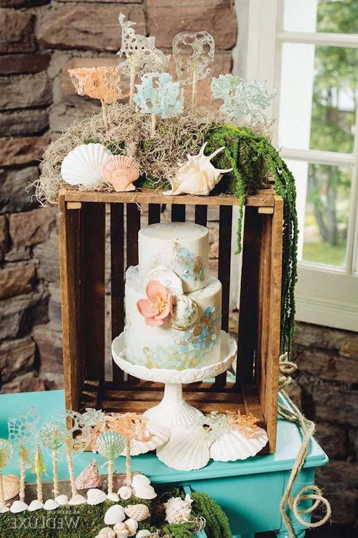 marine-inspired wedding cake, decorated with flowers, near a wooden crate, covered with grey and green moss, and pink and white seashells