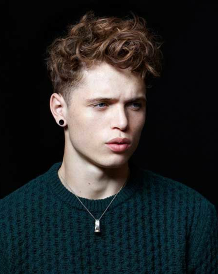angry teen boy, with messy auburn hair, hairstyles for short curly hair, wearing a dark green, textured knitted jumper, and a black stud earring