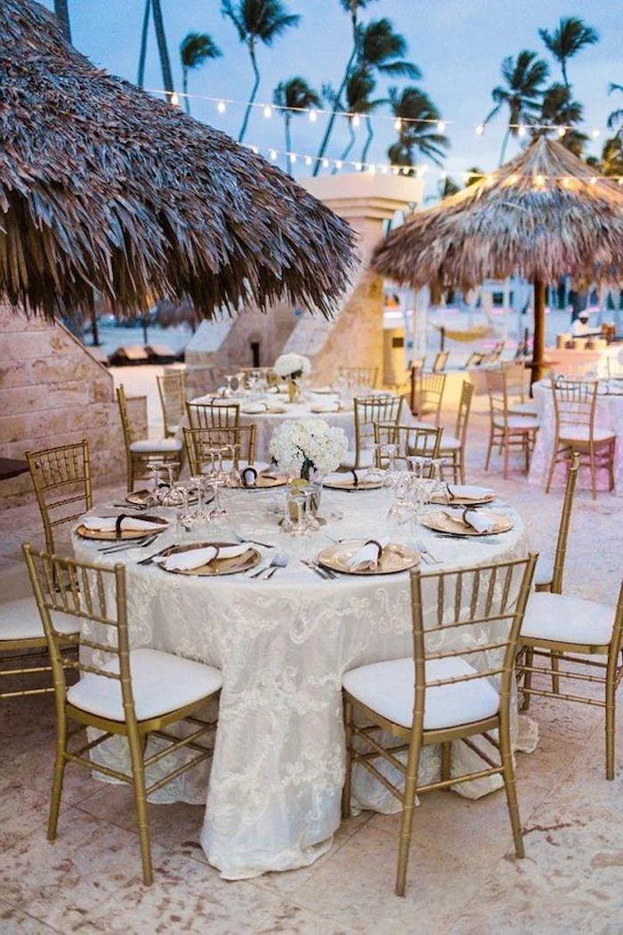 straw-covered sun umbrellas, near several round tables, set for dinner, with lace tablecloths, and gold and white chairs, on a beach with tall palm trees