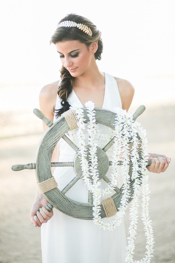 laurel-like silver hair ornament, on brunette woman with messy braid, wedding dresses for beach wedding, wearing plain white gown, and holding a ship's steering wheel, decorated with white garlands