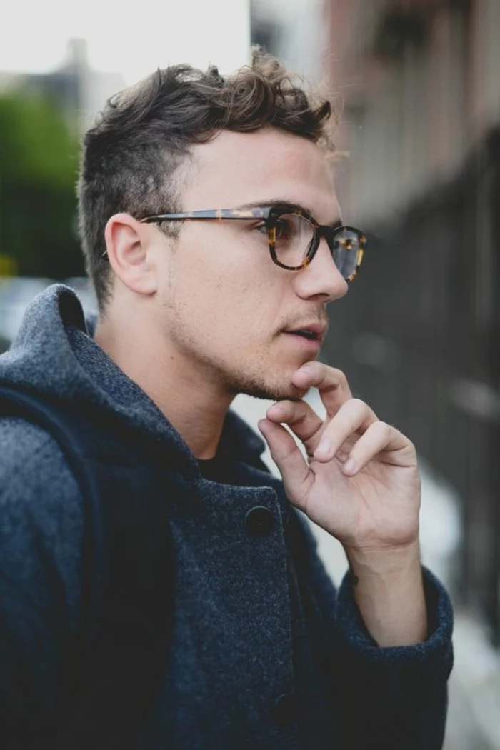 glasses worn by young man, with short curly hair, wearing grey hooded woollen coat, and looking thoughtful, stubble on chin and lip