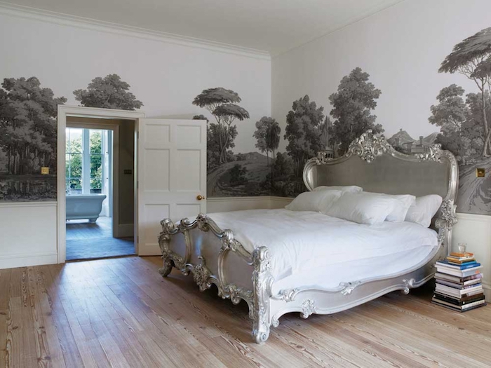 grayscale mural depicting countryside with trees, inside a room with light laminate floor, and a bed with silver baroque style frame, wall art décor in an antique style