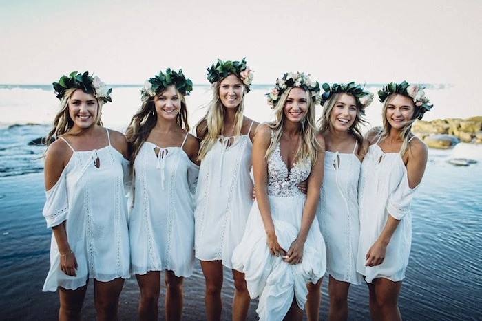 five bridesmaids wearing short, white boho dresses, with cold-shoulder details, and leafy green flower crowns, near a bride with embroidered floaty gown, wedding dresses for beach wedding, sea in the background