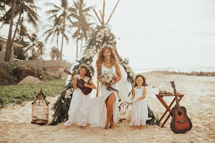 ukulele-playing young girl, next to a smiling bride, in flowy white dress, holding a bouquet, a small girl stands nearby, boho tent decorated with flowers on the beach behind them