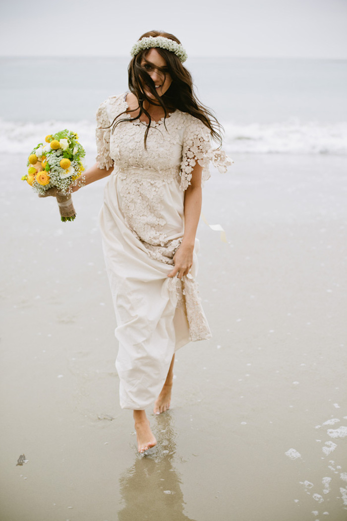 smiling brunette bride, holding a bouquet of white, yellow and green flowers, dressed in off-white embroidered gown, beach wedding dresses, walking barefoot on the shore