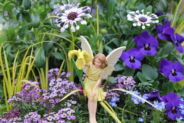 fairy garden ideas, little fairy statuette, with yellow dress, and white wings, holding a yellow flower, in garden with pansies and other purple flowers 
