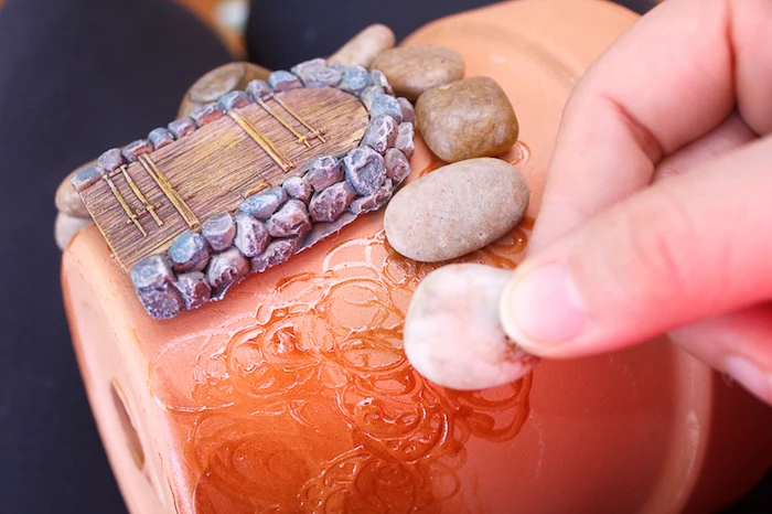 hand sticking a small pale pebble, to an orange ceramic pot, decorated with a tiny painted clay door, fairy garden ideas, beige and brown pebbles