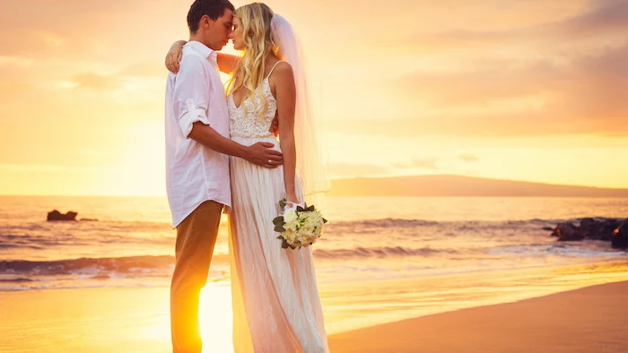 beach wedding, bride in long white boho dress and veil, hugging groom in white shirt, and beige pants, near the sea at sunset
