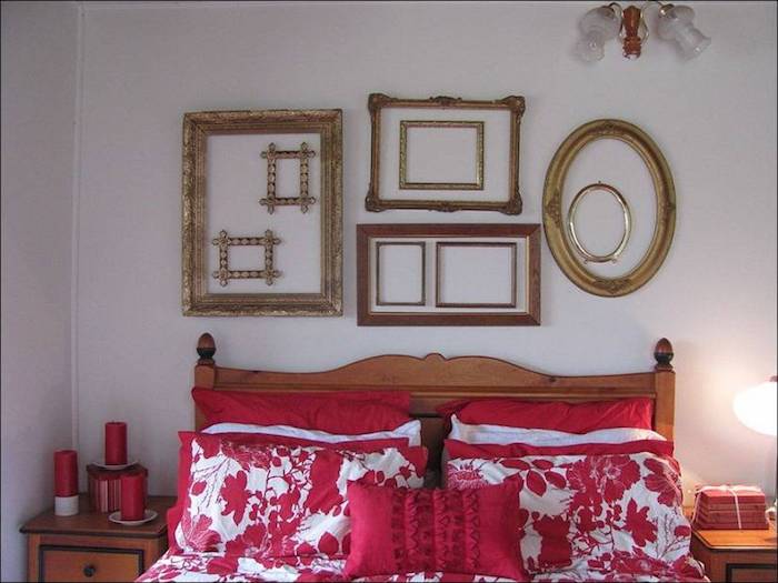 oval and rectangular empty frames, in brown and gold, arranged on a white wall, near brown wooden bed, with red and white pillows and covers 