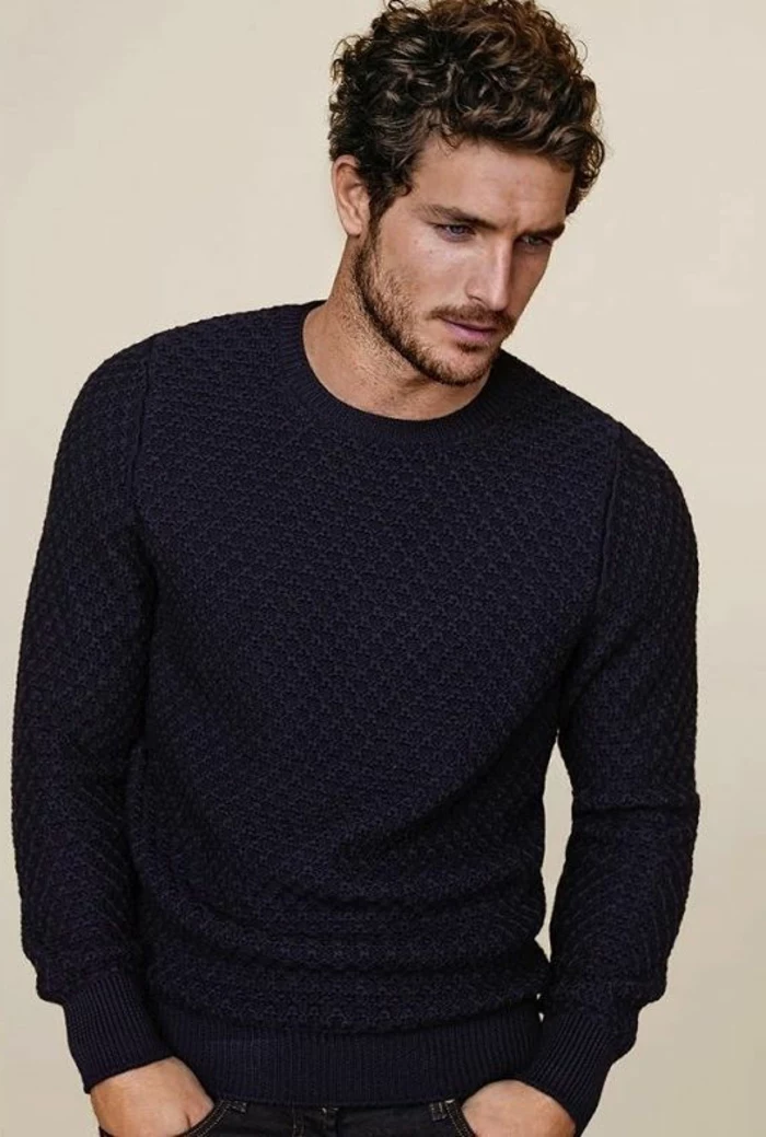 versatile haircut for curly men, worn by a brunette guy, in a textured dark blue knitted jumper, looking to one side, with hands in his pockets